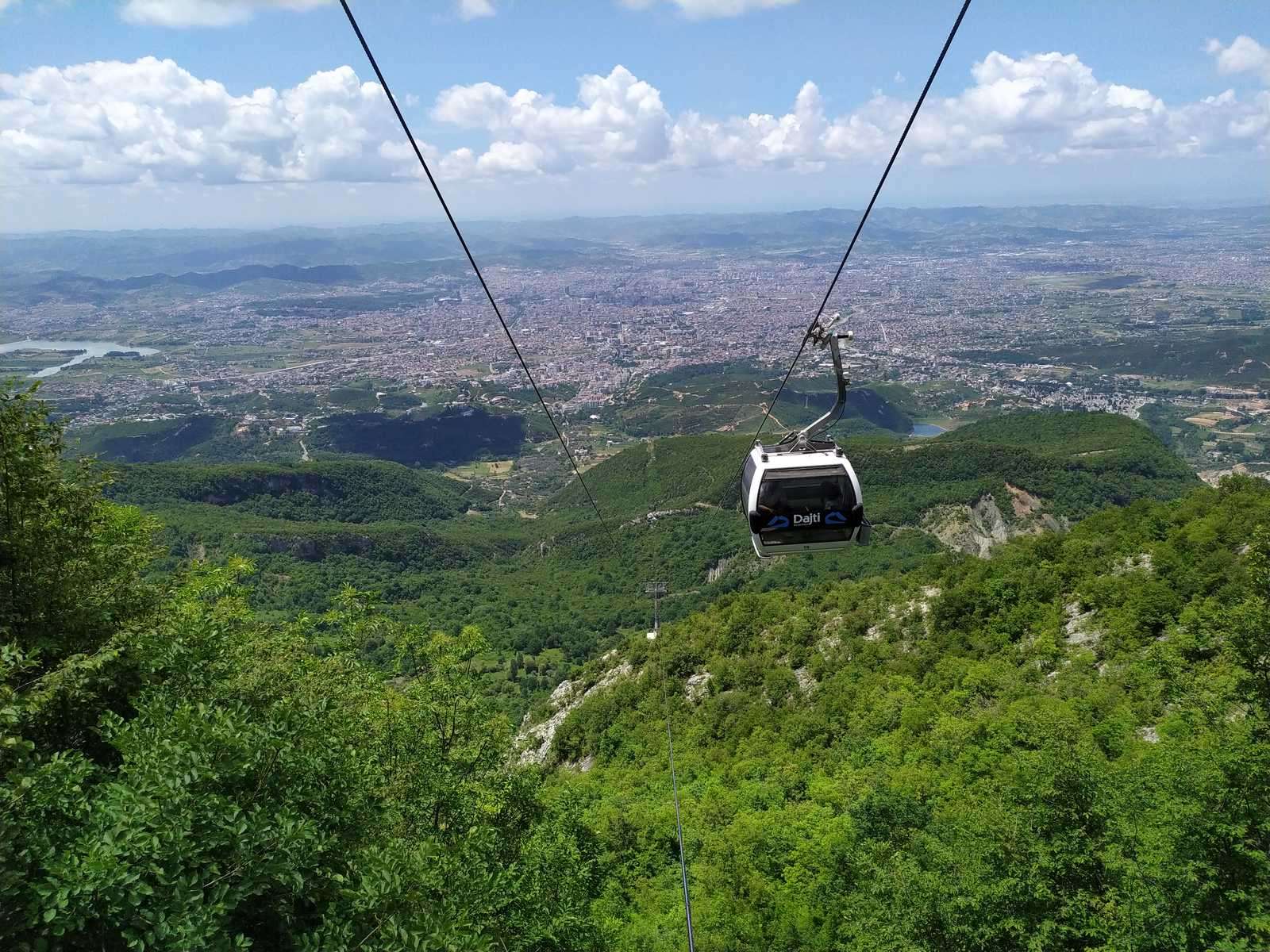 Kabinka lanovky Dajti Ekspres nad zalesněným svahem hory Dajti s panoramatem Tirany v pozadí